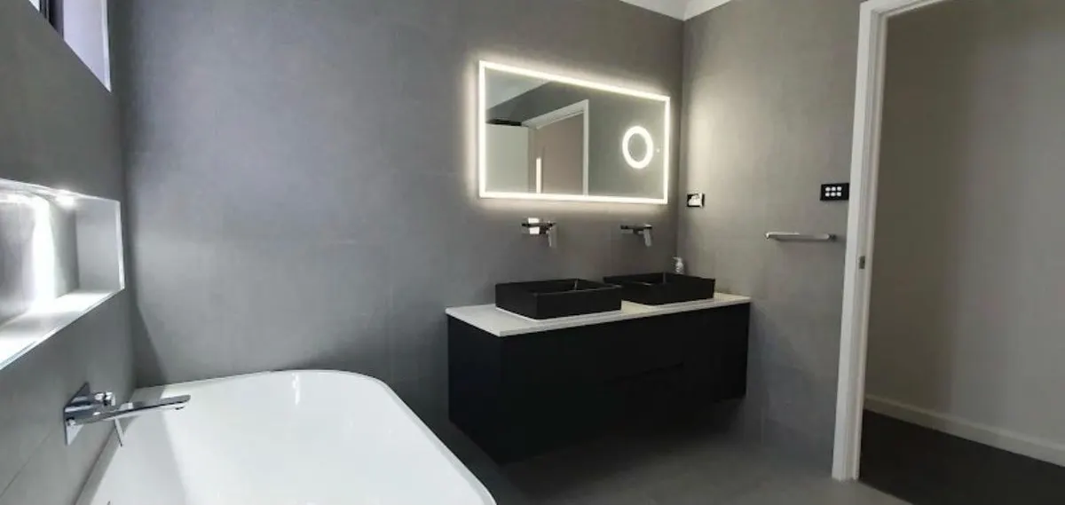 Grey bathroom with double vessel basins on a black floating vanity, backlit mirror and freestanding bath