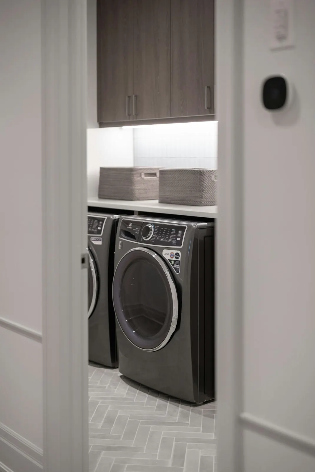 Laundry renovation with front loading washer and dryer, overhead cabinetry and a herringbone tiled floor