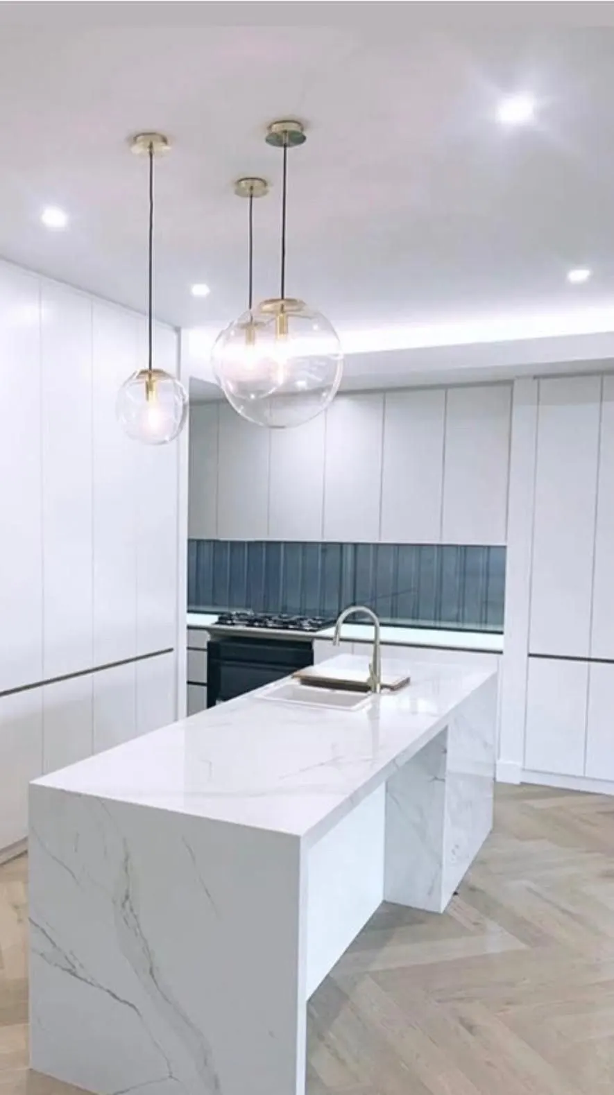 White kitchen with a marble look waterfall island, globe pendant lights and herringbone timber floor